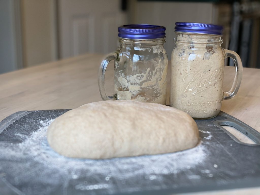 a sourdough loaf unbaked with ball jars behind it full of starter