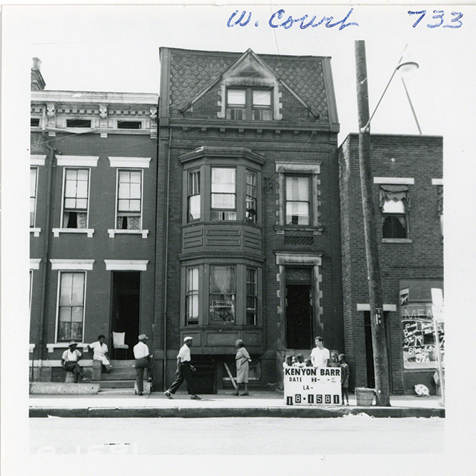 survey photograph depicting row houses with African American men sitting on a stoop and walking down the street. On the right, the surveyor stands with a survey sign for the photograph. The address in the corner reads 733 West Court Street