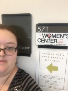 A selfie of the author with the location placard for the University of Cincinnati Women's Center that includes the room number (571 Steger Student Center) and the center's logo