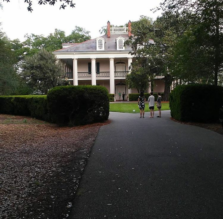 The picture seen is the back of a Greek revivalist architecture southern plantation Big House in southern Louisiana. Around the mainstructure there is a series of landscaping and in the foreground there are the people.