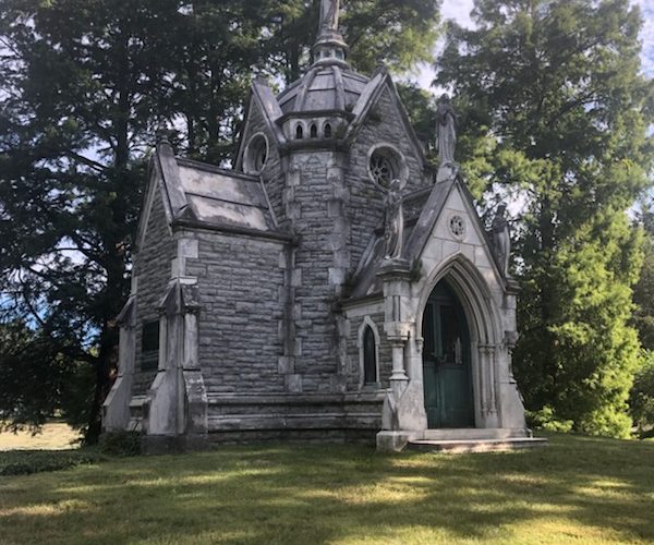 A photo of the gothic style Robinson mausoleum that is surrounded by tall, green trees that cast a shadow on the mausoleum.