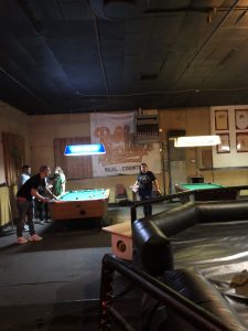 This picture is the inside of Bobby Mackey's Music World. On the left several people are crowded around a pool table. To the right is part of the mechanical bull they have for entertainment. On the back wall is a large poster saying "Bobby Mackey's" in yellow ink. The bar is dimly lit.