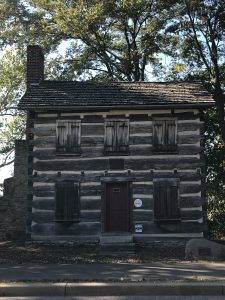 An early 19th century two story log cabin with five windows covered by wooden shutters, a brick chimney, and a red door. It is surrounded and shaded by trees and placed in front of a sidewalk.