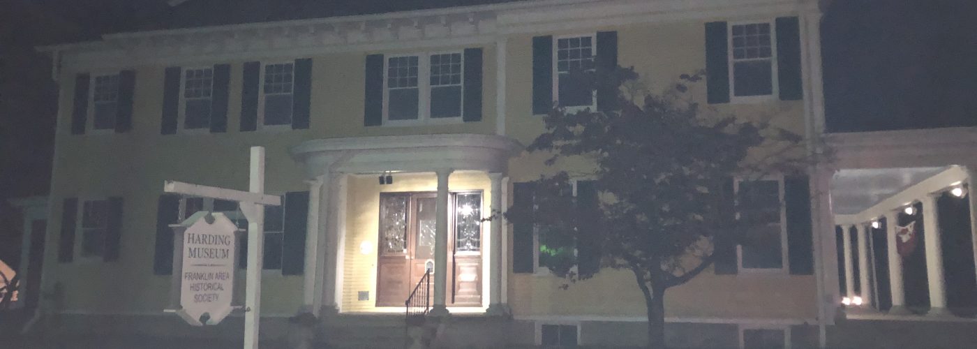 A yellow, two story colonial style house with a lit front porch showing the decorative brown front door. A small tree covers the right side of the house and a white sign reading, "Harding Museum: Franklin Area Historical Society" hangs in the front yard. The image is dark as the photo was taken at night.