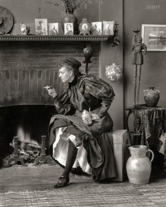 A picture of Frances Benjamin Johnston sitting on a stool. She is in a fancy period dress, but it is pulled up to show off her leg. She is smoking a cigarette, and holds a pitcher of beer in the other hand.