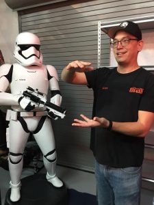 A tall asian man in a Rancho Obi-Wan shirt gestures next to a suit of Storm Trooper armor.