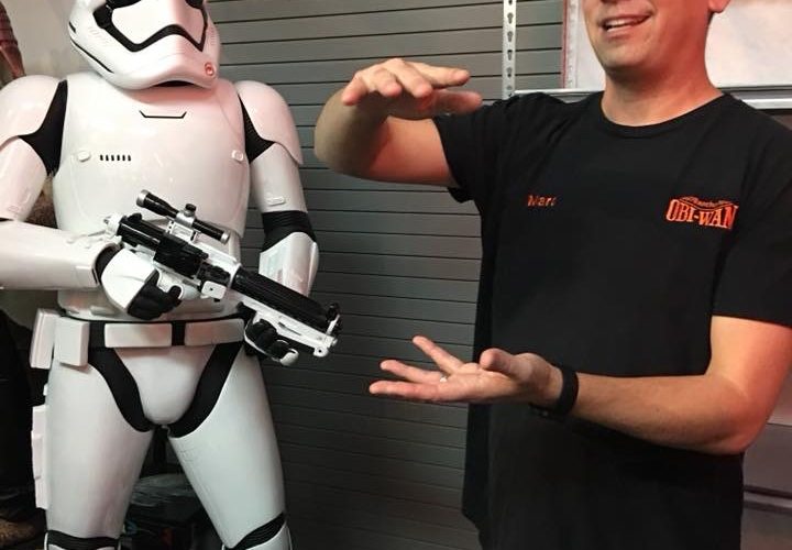 A tall asian man in a Rancho Obi-Wan shirt gestures next to a suit of Storm Trooper armor.