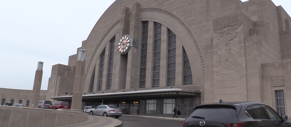 this is a picture of the entrance to the union terminal. the photo is not taken straight on. You see the entrance at an angle. The building is mostly made of tan brick and has a storm windows above the doors. There's also a arc that goes over the windows. In the bottom right is a black car parked.