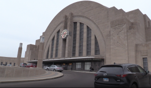 this is a picture of the entrance to the union terminal. the photo is not taken straight on. You see the entrance at an angle. The building is mostly made of tan brick and has a storm windows above the doors. There's also a arc that goes over the windows. In the bottom right is a black car parked.