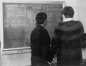 Two men holding hands in front of a blackboard. The board has a table that is showing incoming results for issue 3