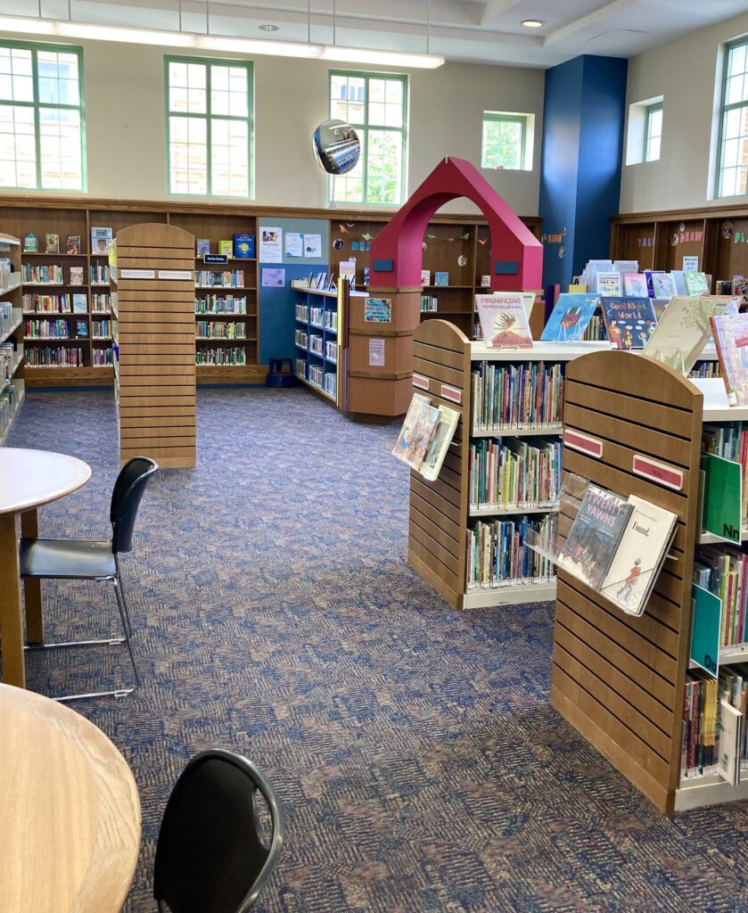 The children's section of a library. Two tables and rows of picture books are visible in the foreground, with more bookshelves in the background.