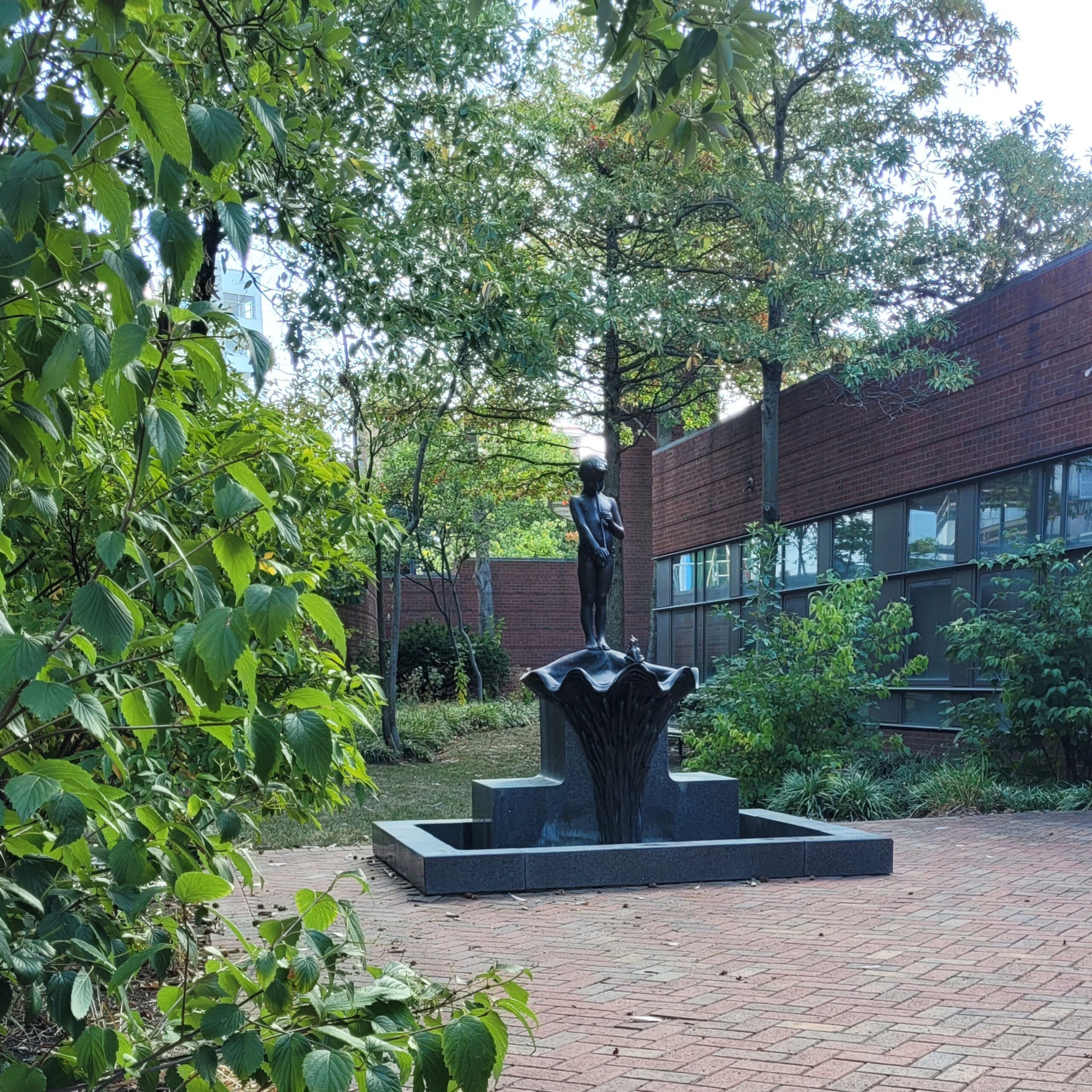 A statue of a nude boy holding a flute and standing on a lily pad surrounded by a greenspace