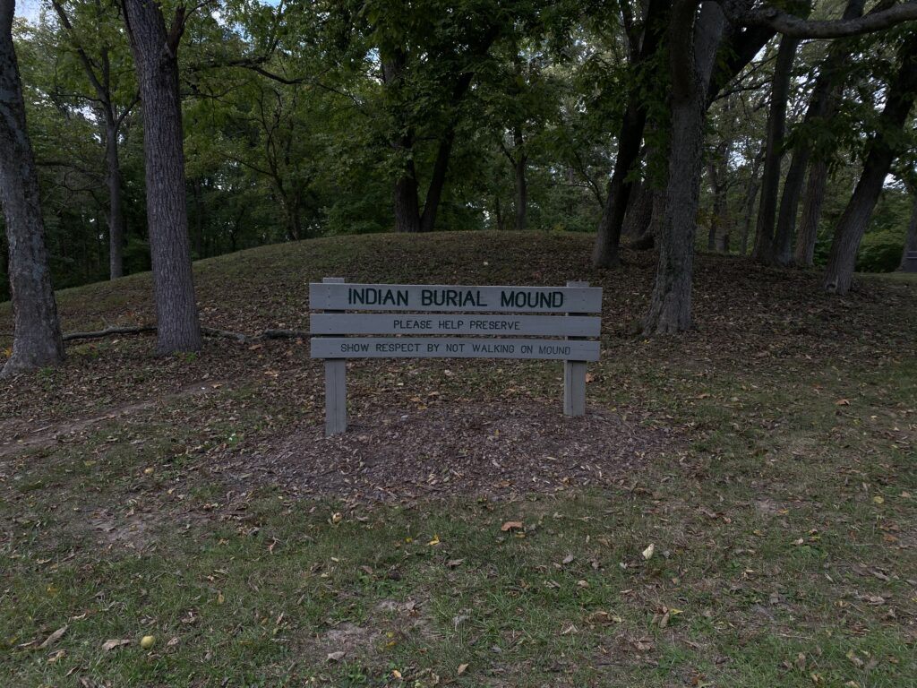 A burial mound marked by wooden signage reading "Indian Burial Mound. Please help preserve. Show respect by not walking on mound."