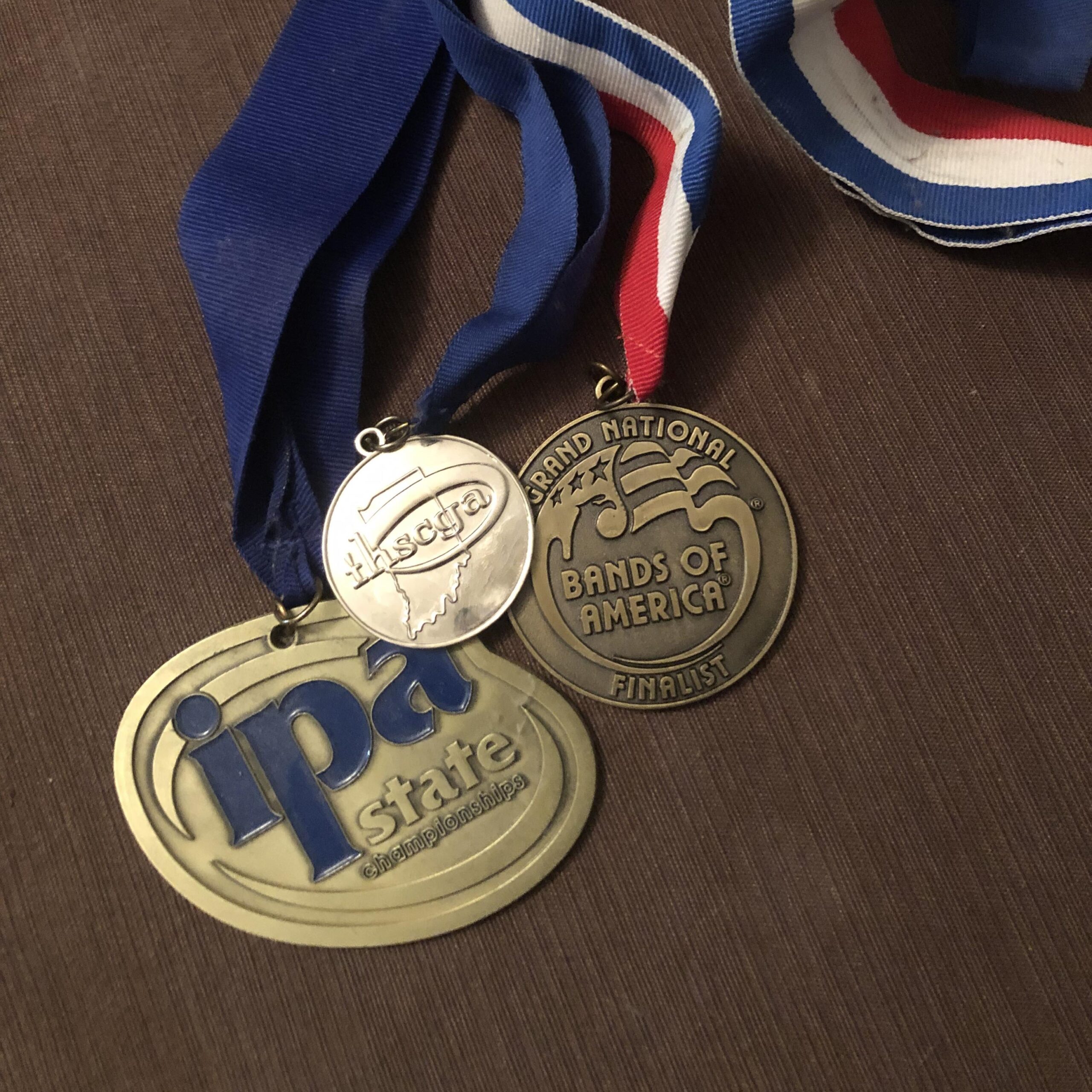 Three different marching band medals on blue or red, white, and blue ribbon against a solid wood background