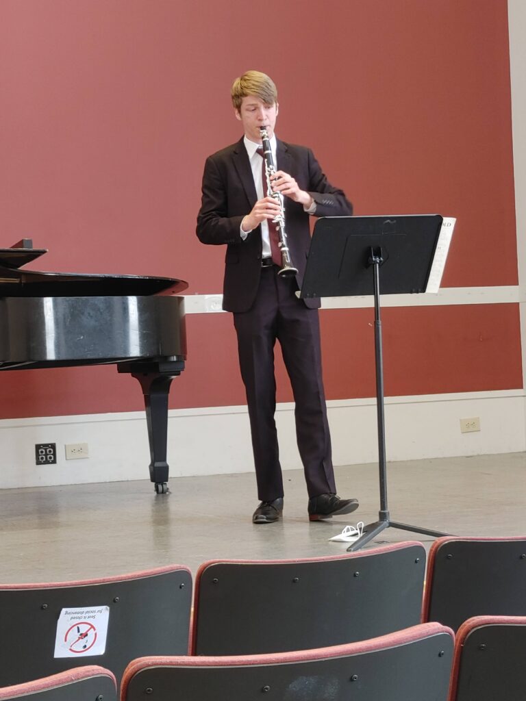 Ian Bunt wearing a suit and tie playing clarinet on stage reading from a music stand.