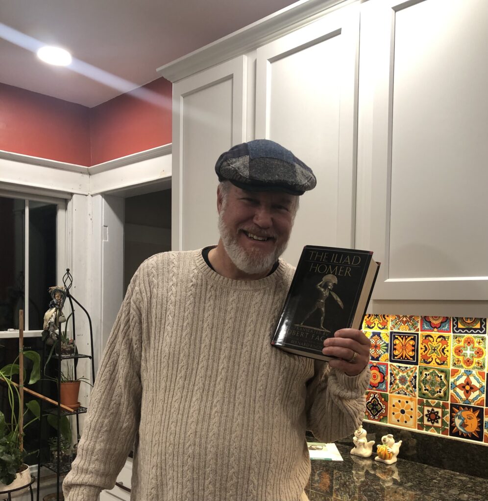 Paul holding the Iliad book up for a photo. Paul is dressed in a white sweater and flat cap. He also sports a white-beard.
