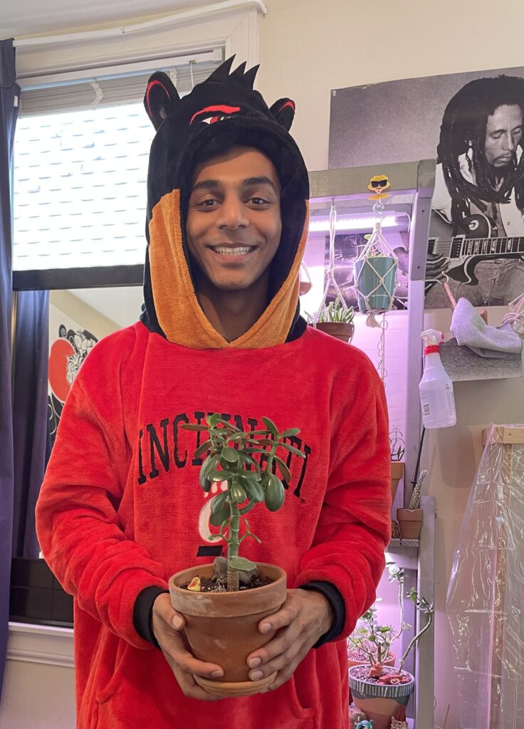 Parth stands with his Jade Plant. Parth is dressed in a red University of Cincinnati sweatshirt.