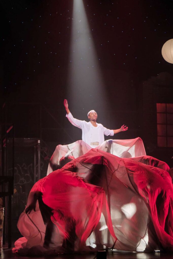 A photograph of a staged production of the musical "Rent" at the moment where the character Angel dies. Billowing red fabric flows around him and a bright white light shines from above.